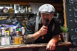 Ein Mann mit Schieberm&uuml;tze und Tattoos spricht ins Mikrofon an einem Gin-Stand. Vor ihm mehrere Flaschen verschiedener Spirituosen.