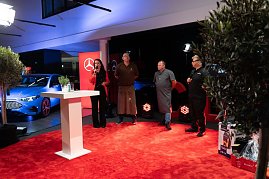 Christina Dost roten Blazer moderiert auf der B&uuml;hne. Neben ihr stehen drei K&ouml;che und Grillmeister in Sch&uuml;rzen und Arbeitskleidung. Im Hintergrund ein blauer Mercedes.