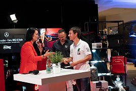 Christina Dost im roten Blazer interviewt einen jungen Mann im wei&szlig;en Mercedes-AMG-Petronas-Shirt. Beide lachen. Dahinter ein Stand mit Mercedes-Merchandise.
