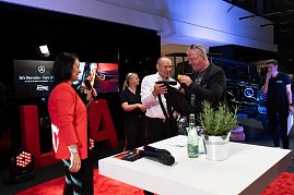 Ein Mann in wei&szlig;em Hemd und ein weiterer in schwarzer Kleidung packen gemeinsam ein Geschenk aus. Links steht Christina Dost im roten Blazer mit Mikrofon.