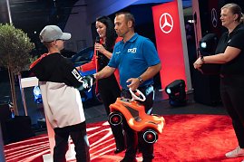 Ein Junge mit Mercedes-Cap bekommt ein rotes Bobbycar von einem Mann im blauen DTZ-Shirt &uuml;berreicht.