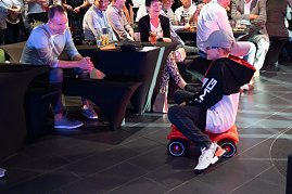 Ein Junge im schwarzen AMG-Hoodie und Mercedes-Cap f&auml;hrt lachend auf einem roten Bobbycar durch den Showroom. Erwachsene G&auml;ste sitzen an Tischen und beobachten ihn am&uuml;siert.