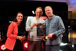 Christina Dost im roten Blazer steht mit Andreas Peter und einem Gewinner auf der B&uuml;hne. Gemeinsam halten sie einen gro&szlig;en Gutschein mit N&uuml;rburgring-Aufdruck in die Kamera.