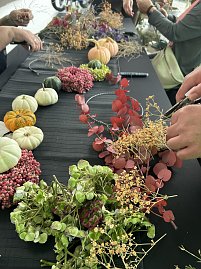 Auf einem Tisch liegen kleine Kürbisse, bunte Beeren und verschiedene Zweige für herbstliche Dekoration. Mehrere Personen gestalten daraus Kränze, einige Hände sind bei der Arbeit zu sehen.
