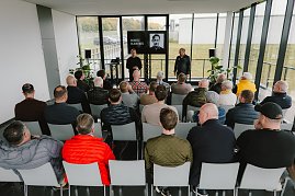 Eine Gruppe von Menschen sitzt in einem modernen, hellen Raum während einer Präsentation. Im Vordergrund sieht man mehrere Männer, die auf Stühlen sitzen und auf eine Bühne blicken. Auf der Bühne stehen zwei Personen: Ein Mann trägt einen schwarzen Pullover und spricht, während der andere in einem schwarzen Outfit steht. Auf einem Bildschirm im Hintergrund wird ein Porträt von Moritz Bleibtreu gezeigt. Der Raum hat große Fenster, durch die man die Natur draußen sieht. Pflanzen stehen in den Ecken des Raumes.