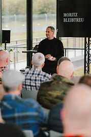 Ein Mann in schwarzem Pullover hält eine Tasse in der Hand und spricht zu einem Publikum. Auf dem Bildschirm hinter ihm ist der Name 'Moritz Bleibtreu' zu sehen. Die Zuschauer im Vordergrund sind nur von hinten zu sehen.