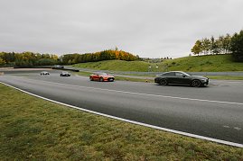 Mehrere Autos fahren auf einer Rennstrecke. Im Vordergrund ist ein schwarzer Mercedes-AMG und ein oranger Mercedes-AMG GT zu sehen. Weitere Fahrzeuge sind in der Ferne sichtbar, während die Strecke von Bäumen gesäumt ist, die herbstliche Farben zeigen. Der Himmel ist bewölkt, und die Strecke führt in eine Kurve.