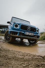 Ein blauer Mercedes-Benz G-Class fährt auf einem matschigen Gelände. Der Blickwinkel zeigt das Fahrzeug aus der Nähe, mit dem Fokus auf die Frontpartie und die markante Kühlergrill. Der Unterboden und die Räder sind mit Schlamm bedeckt, was die Geländetauglichkeit des Fahrzeugs betont. Im Hintergrund ist ein Gebäude unscharf zu sehen.