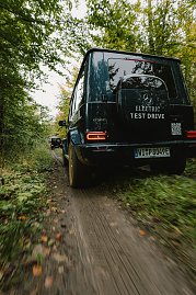 Ein Mercedes-Benz G-Class, Modell G 580, fährt auf einem unbefestigten Waldweg. Das Fahrzeug ist mit dem Aufkleber 'ELECTRIC TEST DRIVE' auf der Heckklappe versehen. Die Reifen sind mit Schlamm bedeckt, und im Hintergrund ist ein weiteres Fahrzeug zu sehen. Der Wald mit grünen Bäumen umgibt den Weg.