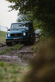 Ein blauer Mercedes-Benz G-Class fährt auf einem unbefestigten, felsigen Weg im Wald. Der Blick ist von der Seite, sodass die Vorderseite des Fahrzeugs und die charakteristischen Scheinwerfer gut sichtbar sind. Im Hintergrund ist ein weiteres Fahrzeug zu sehen. Der Weg ist von Gras und Bäumen gesäumt.