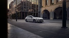 Das Bild zeigt einen weißen Mercedes-Benz CLA Shooting Brake, der auf einer breiten, nahezu leeren Stadtstraße fährt. Moderne Gebäude umgeben die Szene, das Licht wirkt weich und frühmorgendlich. Der Wagen ist seitlich zu sehen und vermittelt Dynamik sowie Eleganz.