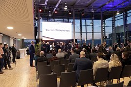 Publikum in einer Messehalle vor einer Bühne mit großer Leinwand Herzlich Willkommen, Gäste im Anzug nehmen Platz.