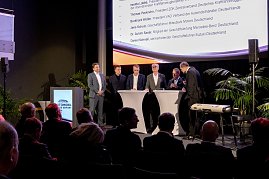 Podiumsrunde auf der Bühne der Automobil Messe Erfurt mit mehreren Männern im Anzug, Publikum im Vordergrund und Teilnehmerliste auf der Leinwand.