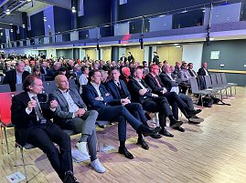 Publikum sitzt in Reihen in einer Messehalle, mehrere Männer im Anzug in der ersten Reihe.