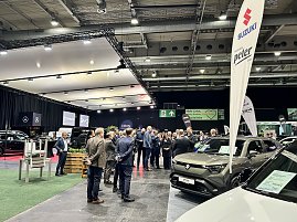 Besuchergruppe auf der Automobilmesse zwischen Messeständen und ausgestellten Autos, rechts ein Suzuki-Stand mit Banner.