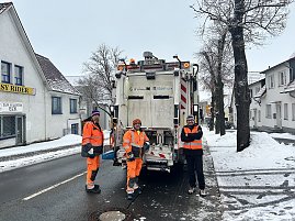 Drei Mitarbeiter der M&uuml;llabfuhr stehen hinter einem M&uuml;llfahrzeug auf einer verschneiten Stra&szlig;e.