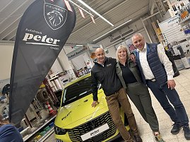 2 M&auml;nner und eine Frau neben einem gelben Peugeot im Baumarkt; daneben schwarze Autohaus Peter Beachflag