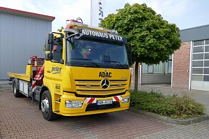 Gelber Abschlepp-LKW des Autohaus Peter im Auftrag des ADAC, Mercedes-Benz, mit der Aufschrift Autohaus Peter auf dem Dachschild und ADAC-Logo an der Front. Das Fahrzeug steht auf einem gepflasterten Platz neben einem Baum und einem grauen Gebäude mit rotem Dach.