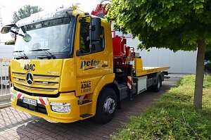 Gelber ADAC-Abschleppwagen mit Mercedes-Stern und der Aufschrift Autohaus Peter an der Seite, geparkt neben einem Baum und Gebäuden, mit rotem Kranaufbau hinten.