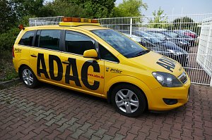 Gelber ADAC-Pannenhilfewagen vom Autohaus Peter, seitlich fotografiert auf einem gepflasterten Parkplatz, mit großem ADAC-Schriftzug und orangenem Warnlicht auf dem Dach. Im Hintergrund stehen weitere Fahrzeuge hinter einem weißen Zaun.