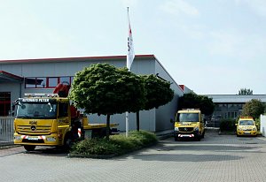 Drei gelbe ADAC-Fahrzeuge des Autohaus Peter, darunter ein Abschlepp-LKW, ein Transporter und ein PKW, stehen vor einem grauen Gebäude mit roten Fensterrahmen. Davor stehen zwei Bäume und ein Fahnenmast mit Fahne. Der Himmel ist leicht bewölkt.
