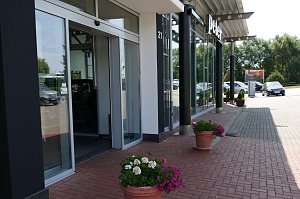  Eingangsbereich eines Autohauses der Peter Gruppe in Duderstadt, mit gro&szlig;en Glasfassaden, schwarzer Beschilderung und Blumen in T&ouml;pfen entlang des gepflasterten Weges; rechts stehen geparkte Autos, im Hintergrund B&auml;ume und ein Parkplatz.