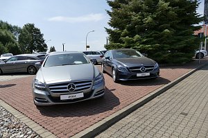 Zwei Mercedes-Benz Fahrzeuge stehen nebeneinander auf einem gepflasterten Parkplatz. Links ein silberner Mercedes und rechts ein dunkler Mercedes. Im Hintergrund weitere Fahrzeuge und gro&szlig;e gr&uuml;ne Tannenb&auml;ume. 