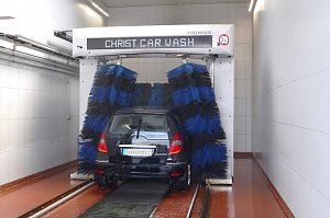 Auto in einer automatischen Waschanlage. Blaue und schwarze B&uuml;rsten reinigen das schwarze Fahrzeug. Die Umgebung ist hell und gekachelt, mit rutschfestem Boden.