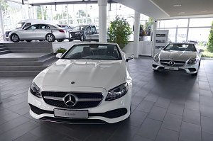 Heller, moderner Mercedes-Benz-Showroom mit gro&szlig;en Glasfronten. Im Vordergrund ein wei&szlig;es E-Klasse Cabriolet, dahinter ein silbernes Cabriolet. Auf einer erh&ouml;hten Fl&auml;che stehen weitere Mercedes-Fahrzeuge. Viel Tageslicht, freundliche und offene Atmosph&auml;re.