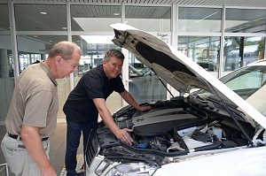 Zwei M&auml;nner stehen vor einem wei&szlig;en Auto mit ge&ouml;ffneter Motorhaube. Einer der M&auml;nner tr&auml;gt ein schwarzes Polohemd und zeigt dem anderen den Motorraum. Beide wirken konzentriert und interessiert.
