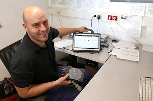 Ein freundlicher Mitarbeiter im schwarzen Polohemd sitzt an einem Schreibtisch in einem B&uuml;ro. Er h&auml;lt ein Diagnoseger&auml;t in der Hand und zeigt l&auml;chelnd auf ein Tablet mit einer Fahrzeug-Software.