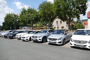 Reihe von Mercedes-Benz Gebrauchtwagen in verschiedenen Farben, geparkt auf einem Autohaus-Au&szlig;enplatz. Im Hintergrund stehen B&auml;ume, ein Wohnhaus mit rotem Dach und weitere H&auml;user, blauer Himmel mit einigen Wolken.