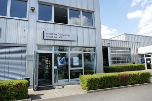 Eingangsbereich eines Autohauses mit Glasfront. &Uuml;ber der T&uuml;r h&auml;ngt ein Schild mit der Aufschrift �Annahme Transporter, Annahme LKW�. Rechts ist eine Werkstatthalle mit gro&szlig;en Toren sichtbar, davor gepflegte Hecken und ein gepflasterter Weg.