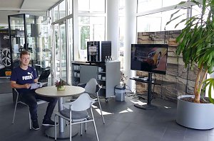 Heller Wartebereich im Mercedes-Autohaus mit Glasfront. Ein junger Mann sitzt an einem runden Tisch und liest eine Brosch&uuml;re. Neben ihm stehen weitere St&uuml;hle, ein kleiner Tisch mit einer Pflanze, ein Kaffeeautomat mit Tassenregal und ein gro&szlig;er Fernseher. Rechts im Bild eine gro&szlig;e gr&uuml;ne Pflanze in einem grauen Topf.