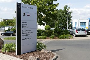 Gro&szlig;es wei&szlig;es Schild auf dem Gel&auml;nde eines Mercedes-Benz Autohauses. Auf dem Schild stehen Logos von Mercedes-Benz und Smart sowie Hinweise auf angebotene Leistungen: Verkauf, Service, OMNIplus, Teile/Zubeh&ouml;r, Gebrauchtwagen, Junge Sterne und Van ProCenter. Im Hintergrund sieht man geparkte Autos, gr&uuml;ne Str&auml;ucher und Geb&auml;ude.