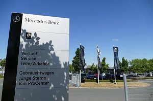 Großes weißes Mercedes-Benz Hinweisschild mit Smart-Logo, Beschriftung für Verkauf, Service, Teile/Zubehör, Gebrauchtwagen, Junge Sterne und Van ProCenter; im Hintergrund wehen mehrere Mercedes- und Smart-Flaggen, Bäume und geparkte Autos unter blauem Himmel.