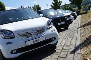 Nahaufnahme einer Reihe von Smart-Autos in Weiß und Schwarz, schräg geparkt auf einem gepflasterten Parkplatz vor dem Autohaus Peter in Northeim, sonniger Tag mit Bäumen und Hügeln im Hintergrund.