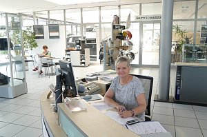 Freundliche Mitarbeiterin sitzt an einem Empfangstresen im Autohaus, vor ihr ein Computer, Telefon und Unterlagen. Sie l&auml;chelt in die Kamera. Im Hintergrund sitzt eine weitere Frau an einem Tisch und liest. Helle, moderne Empfangshalle mit Glasw&auml;nden, Pflanzen und Schildern, auf einem Glast&uuml;r-Bereich steht �Serviceannahme�.