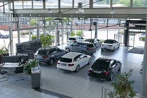 Großer, heller Showroom des Autohauses mit großen Glasfronten. Mehrere Mercedes-Benz Fahrzeuge in verschiedenen Farben (schwarz, silber, weiß) stehen ausgestellt. Links Bürotische mit Pflanzen und Unterlagen. Draußen sind weitere Autos und Grünflächen sichtbar. Die Atmosphäre wirkt modern und einladend.