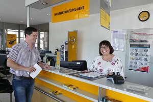 Ein Mann und eine Frau stehen am Empfangstresen mit der Aufschrift INFORMATION / OPEL-RENT. Die Frau hinter dem gelb-wei&szlig;en Tresen tr&auml;gt eine Brille und ein gebl&uuml;mtes Oberteil, sie l&auml;chelt. Der Mann vor dem Tresen h&auml;lt Unterlagen in der Hand und l&auml;chelt ebenfalls. Im Hintergrund h&auml;ngen Opel-Plakate, es ist ein gelbes Opel-Logo an der Wand sichtbar. Der Empfangsbereich wirkt hell und freundlich.