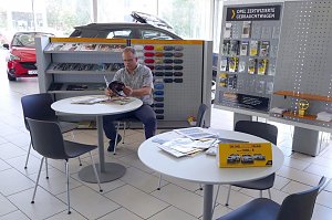 Ein Mann sitzt in einer hellen Opel-Ausstellungshalle an einem Tisch und liest eine Brosch&uuml;re. Im Hintergrund sind Regale mit Prospekten, Farbmustern und Zubeh&ouml;r zu sehen. Rechts befindet sich eine Tafel mit dem Hinweis Opel zertifizierte Gebrauchtwagen. Auf den Tischen liegen Zeitschriften und Flyer. Es sind mehrere schwarze St&uuml;hle um die Tische angeordnet. 