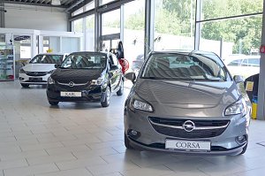  In einem hellen Autohaus-Ausstellungsraum stehen drei Opel-Fahrzeuge. Im Vordergrund ein grauer Opel Corsa, daneben ein schwarzer Opel Karl und im Hintergrund ein wei&szlig;es Fahrzeug. Der Raum hat gro&szlig;e Fenster, durch die viel Tageslicht einf&auml;llt. Im Hintergrund sind Glaskabinen und Vitrinen mit Zubeh&ouml;r zu sehen.