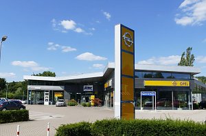 Au&szlig;enansicht eines Opel-Autohauses in Osterode. Das moderne Geb&auml;ude hat eine gro&szlig;e Glasfront und ein markantes, gelb-blaues Opel-Logo auf einem hohen Pylon im Vordergrund. Vor dem Autohaus sind mehrere Fahrzeuge geparkt. Der Himmel ist blau mit einigen kleinen Wolken, wodurch die Szene freundlich und einladend wirkt.