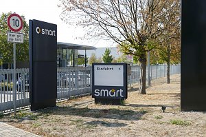 Einfahrtbereich eines Smart-Autohauses mit Wegweiser, Geschwindigkeitsbegrenzungsschild (10 km/h) und B&auml;umen, im Hintergrund das Autohaus.