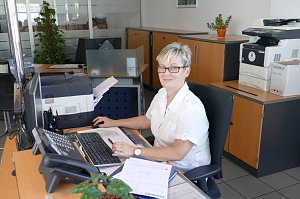Freundliche Mitarbeiterin sitzt an einem Schreibtisch im Autohaus-B&uuml;ro, vor einem Computer mit Unterlagen, Drucker und Schr&auml;nken im Hintergrund