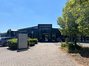 Der moderne Eingangsbereich des Autohauses mit gro&szlig;en Fenstern und der Aufschrift Mercedes-Benz. Im Vordergrund steht ein Wegweiser mit den Bereichen: Verkauf, Service und Teile/Zubeh&ouml;r. Rechts steht ein Baum mit gr&uuml;nen Bl&auml;ttern.