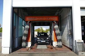 Ein grauer Smart fährt gerade in eine Autowaschanlage mit orangefarbener Beschriftung ‚Christ CAR WASH‘. Links und rechts hängen graue Bürsten.