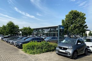 Außenansicht des Mercedes-Benz Autohauses Peter in Dessau. Im Vordergrund steht ein grauer Smart mit dem Schild ‚Gebrauchtwagen‘. Daneben viele schwarze und silberne Mercedes-Benz Fahrzeuge in einer Reihe auf dem Parkplatz. Im Hintergrund modernes Glasgebäude mit großen Fenstern, Bäumen und blauem Himmel mit leichten Wolken.