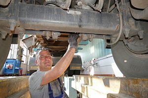 Ein Werkstattmitarbeiter steht unter einem großen LKW und arbeitet an dessen Unterseite. Er trägt graues T-Shirt, schaut in die Kamera und lächelt. Im Hintergrund ist die Werkstatt mit Werkzeugen zu sehen.