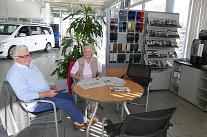 Zwei ältere Kunden sitzen im Wartebereich eines Mercedes-Autohauses an einem Tisch mit Zeitungen, im Hintergrund ein weißer Van, Prospektständer und Farbmusterwand.